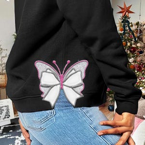 Könnte beinhalten: Schwarzer Hoodie mit einem weißen Schmetterling mit rosa Akzenten, der auf der Rückseite gestickt ist. Der Schmetterling hat eine Schleifenform in der Mitte.