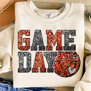 Puede incluir: Una sudadera beige con las palabras "GAME DAY" en letras de purpurina naranja y negra. Una pelota de baloncesto con un corazón está en el centro del diseño.