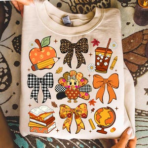 May include: Beige sweatshirt with a colorful fall-themed graphic. The design includes an apple, pencil, bows, turkey, coffee, books, globe, and fall leaves. The graphic is in shades of orange, brown, black, and white.