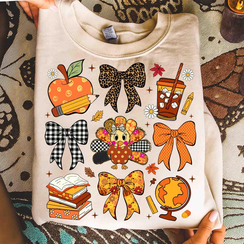 May include: Beige sweatshirt with a colorful fall-themed graphic. The design includes an apple, pencil, bows, turkey, coffee, books, globe, and fall leaves. The graphic is in shades of orange, brown, black, and white.
