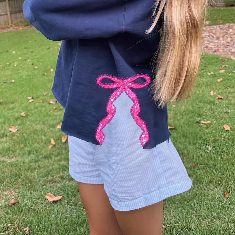Puede incluir: Una sudadera azul marino con un lazo de lentejuelas rosa en la manga. La sudadera se combina con pantalones cortos de rayas azules.