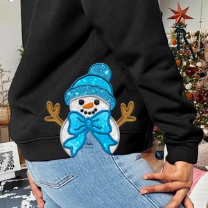 May include: Black sweatshirt with a snowman design. The snowman has a blue glitter hat and bow, with brown stick arms and a carrot nose. The design is centered on the sweatshirt, creating a festive winter theme.