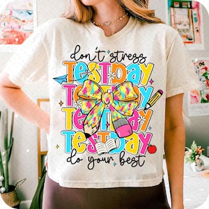 May include: White t-shirt with a colorful graphic that says "Don't stress test day do your best". The graphic features a bow, pencils, and a book.