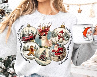 Op de afbeelding: Een wit sweatshirt met een kerstthema ontwerp met zes ornamenten met verschillende kerstscènes, waaronder een hert, een sneeuwpop, een peperkoekman, een kerstboom en de kerstman. De tekst "It's the most wonderful time of the year" wordt twee keer rond de ornamenten herhaald.