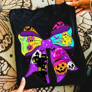 May include: Black t-shirt featuring a colorful Halloween bow design. The bow includes a haunted house, jack-o'-lantern, ghost, and skull. The bow is purple, yellow, orange, and blue. The shirt is held by two hands.