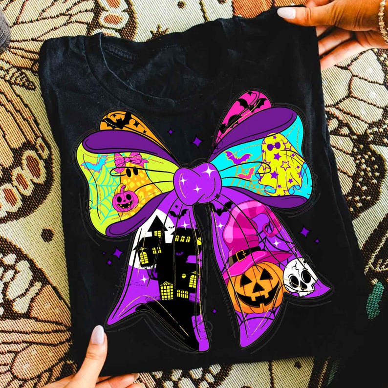May include: Black t-shirt featuring a colorful Halloween bow design. The bow includes a haunted house, jack-o'-lantern, ghost, and skull. The bow is purple, yellow, orange, and blue. The shirt is held by two hands.