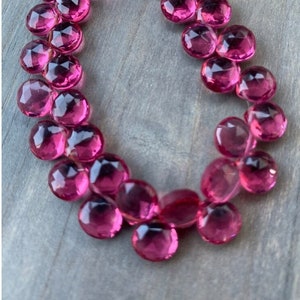Pode incluir: Fio de pedras preciosas rosa choque, redondas e facetadas. As contas são enfiadas, criando uma forma curva. As pedras preciosas têm uma superfície polida e refletora, sobre um fundo de madeira cinza claro.