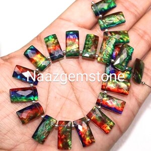Può includere: Un filo di pietre preziose rettangolari e iridescenti nelle tonalità del verde, rosso e blu. Le pietre sono infilate con piccole perle d'argento. Il testo "Naazgemstone" è visibile in bianco.