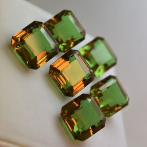 May include: Six emerald green and orange gemstones with a square cut. The gemstones are arranged in a staggered pattern on a white surface.