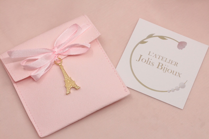 May include: A pink velvet jewelry pouch with a gold Eiffel Tower charm and a pink satin ribbon. The pouch has a white card with the text "L'Atelier Jolis Bijoux".