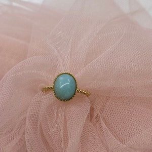 May include: A gold ring with a light blue oval gemstone set in a bezel setting. The ring is on a pink tulle fabric.