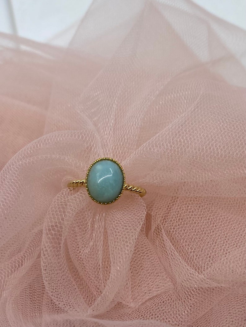 May include: A gold ring with a light blue oval gemstone set in a bezel setting. The ring is on a pink tulle fabric.