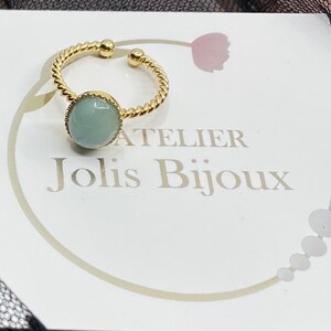 May include: A gold ring with a twisted band and a light blue gemstone. The ring is on a white card with the text "Atelier Jolis Bijoux".