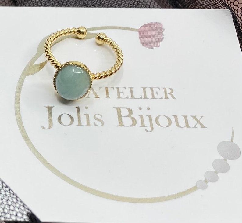 May include: A gold ring with a twisted band and a light blue gemstone. The ring is on a white card with the text "Atelier Jolis Bijoux".