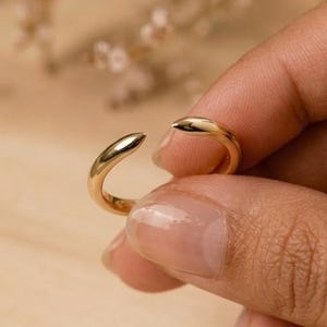 Op de afbeelding: Een goudkleurige open ring. De ring heeft een glad, gepolijst oppervlak en een minimalistisch ontwerp. De ring wordt vastgehouden door de hand van een persoon, met een houten oppervlak op de achtergrond. De ring is een sieraad.