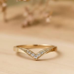 以下が含まれることがあります： 上部の曲線に沿って小さな透明な宝石がセットされた、金色のV字型のリング。リングは明るい色の木製の表面にあり、背景にはぼやけた花の要素があります。