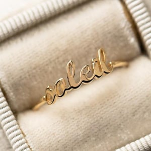 May include: A gold ring inscribed with the word "soleil" in cursive lettering. The ring is presented in a grey velvet ring box. "Soleil" is French for "sun".