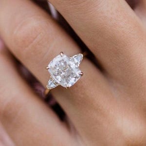 Op de afbeelding: Een close-up van een ring met een grote, kussen geslepen diamant als middelste steen en twee kleinere, peervormige diamanten aan weerszijden. De ring is gezet in een gouden band.