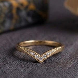 May include: A gold chevron-shaped ring featuring a series of small, clear, round-cut stones set along the V-shaped band. The metal band displays a polished finish with delicate milgrain detailing along the edges of the stone setting.