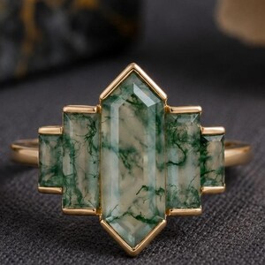 May include: A gold ring featuring a multi-stone arrangement of moss agate. The center stone is a large, elongated hexagon, flanked by two smaller rectangular stones on each side, all set in a gold frame with a geometric, stepped design.