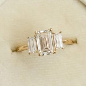 Könnte beinhalten: Ein goldener Ring mit drei rechteckigen, klaren Edelsteinen. Der Mittelstein ist größer als die beiden Seitensteine. Der Ring befindet sich in einer Samt-Ringbox, vor einem weißen Satintuch.