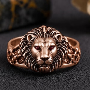 May include: A rose gold-toned ring featuring a detailed lion head centerpiece with small, deep red gemstone eyes. The band of the ring displays an intricate, woven or braided metal pattern that extends from the sides of the lion head design.