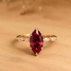 May include: A gold ring with a marquise-cut, deep red gemstone as the focal point. The band features small, clear accent stones. The ring is set against a light wooden surface, with soft, blurred background elements.