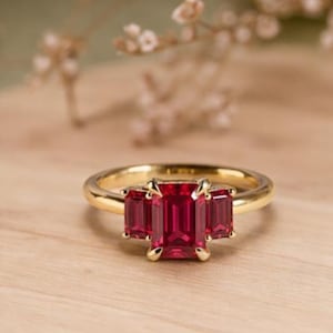 May include: A gold ring featuring three rectangular, vibrant red gemstones. The ring is set on a light wooden surface, with soft, blurred floral elements in the background. The ring's design is elegant and eye-catching.
