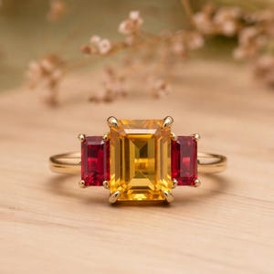 May include: A gold ring featuring a large, rectangular yellow gemstone flanked by two smaller, rectangular red gemstones. The ring is set on a light-colored wooden surface, with a blurred background of greenery and dried flowers.