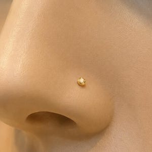 Puede incluir: Un pequeño piercing de nariz redondo en tono dorado con una única piedra preciosa transparente y facetada en el centro. La joya está colocada en el lateral de una nariz, mostrando un diseño minimalista con un acabado metálico pulido.