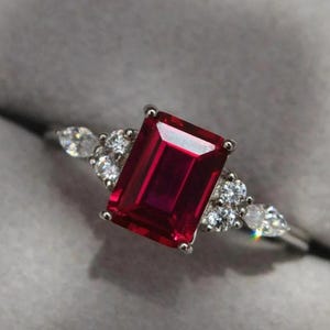 Może przedstawiać: Srebrny pierścionek z dużym, prostokątnym, głęboko czerwonym kamieniem centralnym. Mniejsze, przezroczyste kamienie flankują główny kamień. Pierścionek jest umieszczony na szarym tle.