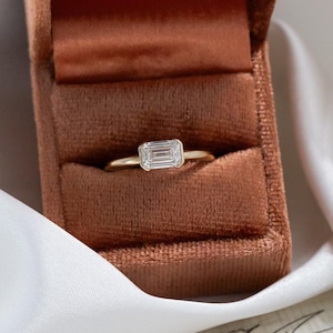 May include: A gold ring featuring a rectangular emerald-cut diamond, presented in a velvet ring box. The ring is set against a white cloth and a surface with script writing.