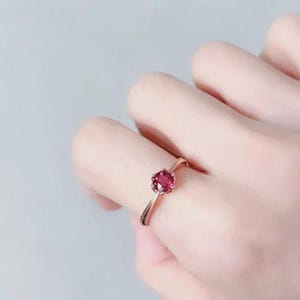 Puede incluir: Un anillo de oro rosa con una gema redonda de color rojo intenso. La banda del anillo tiene un diseño delicado y texturizado. La gema está engastada en un engaste clásico de garras. El anillo se muestra en un dedo.