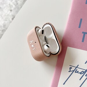 May include: A pink leather case for wireless earbuds with a white star design. The case is open, revealing the white earbuds inside.