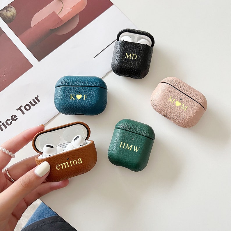 May include: Five different Airpod cases with personalised initials and a small heart. The cases are in brown, black, teal, pink, and green leather.