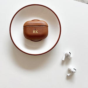 May include: A brown leather case for wireless earbuds with the initials "RK" embossed on the front. The case is sitting on a white plate.