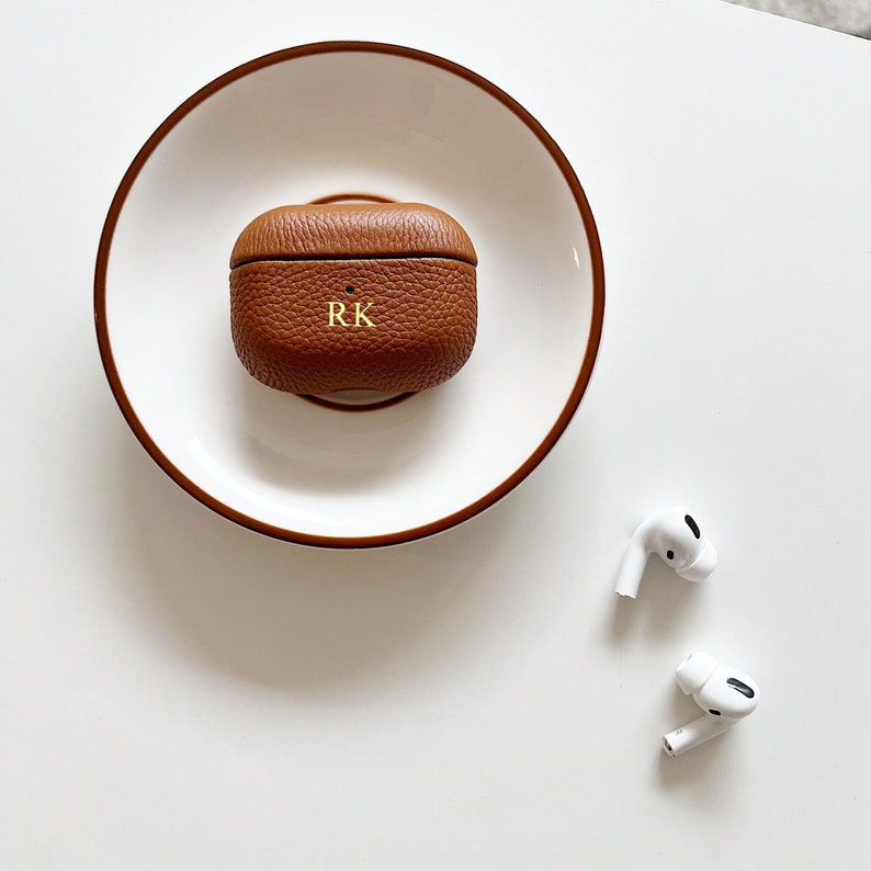 May include: A brown leather case for wireless earbuds with the initials "RK" embossed on the front. The case is sitting on a white plate.