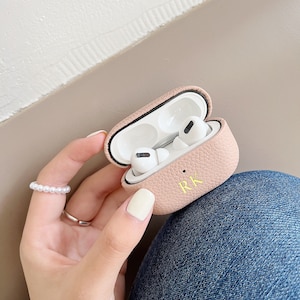 May include: A pink leather case for AirPods with the initials "RK" embossed in gold. The case is open, revealing the white AirPods inside.