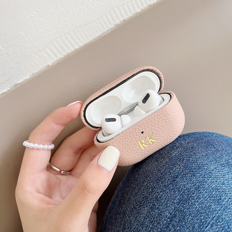 May include: A pink leather case for AirPods with the initials "RK" embossed in gold. The case is open, revealing the white AirPods inside.