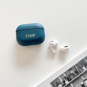 May include: A teal blue leather case with the word "rose" embossed on the front. Two white wireless earbuds are next to the case.