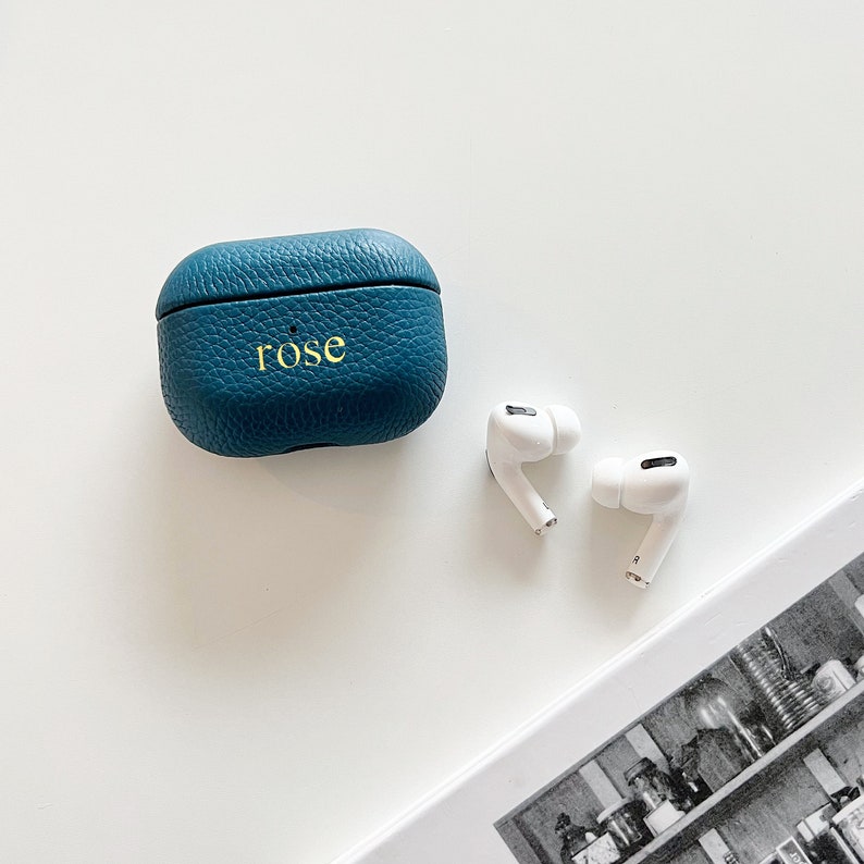 May include: A teal blue leather case with the word "rose" embossed on the front. Two white wireless earbuds are next to the case.