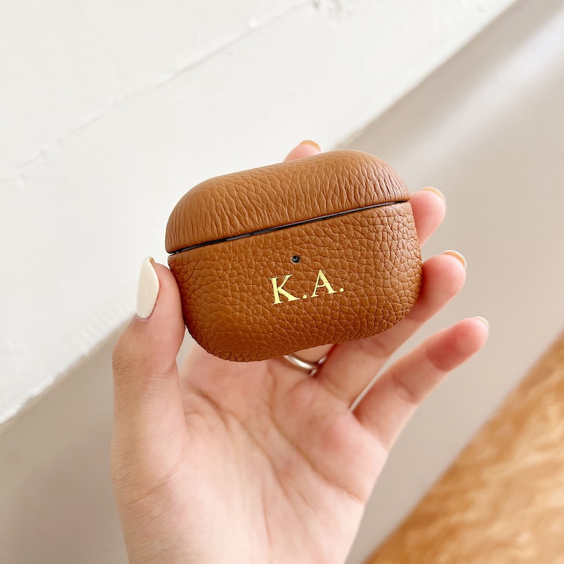 May include: A brown leather Airpods case with the initials "K.A." embossed in gold lettering.