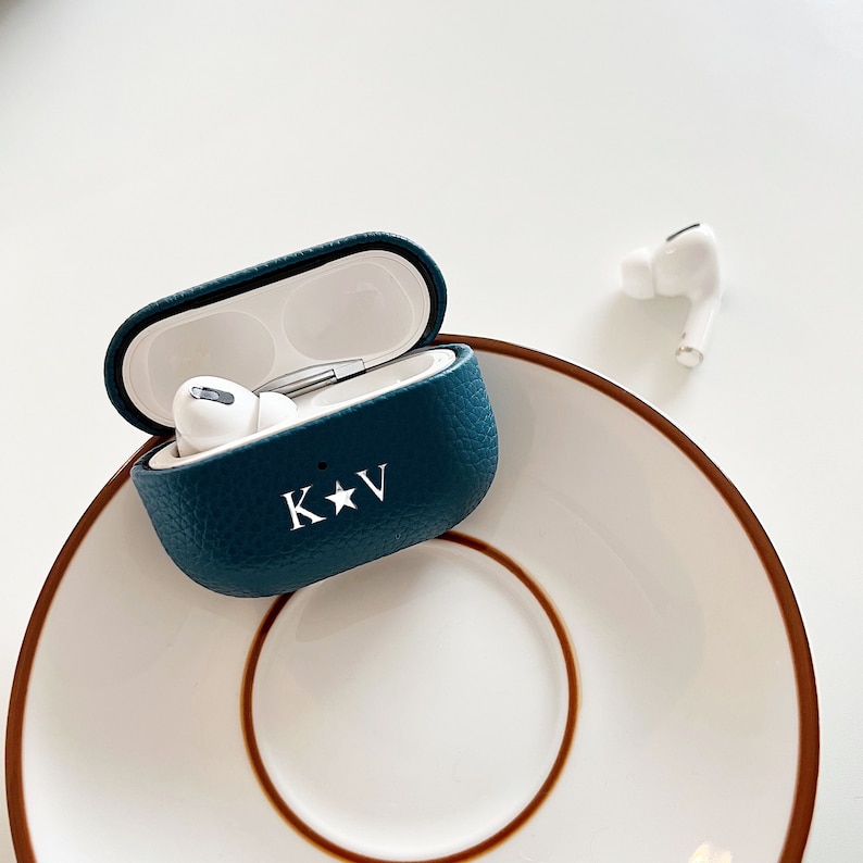 May include: A teal blue leather case for wireless earbuds with the initials "K*V" embossed on the front. The case is open and the earbuds are visible inside.
