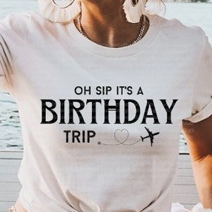 以下が含まれることがあります： 黒い文字で「Oh Sip It's A Birthday Trip」と書かれた白いTシャツ。ハートと飛行機のグラフィックが描かれている。