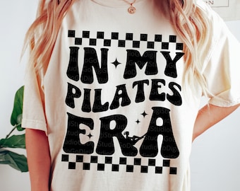 May include: A white t-shirt with a black graphic that says "In My Pilates Era" with a silhouette of a person doing pilates. The graphic is surrounded by a black and white checkered border.