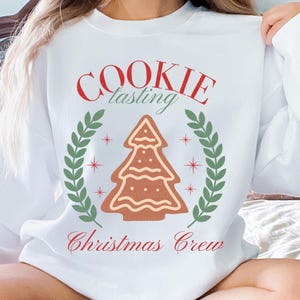Könnte beinhalten: Weißes Sweatshirt mit dem roten Schriftzug "COOKIE tasting Christmas Crew". Ein Lebkuchen-Weihnachtsbaum befindet sich in der Mitte eines grünen Kranzes. Rote Sterne akzentuieren den Kranz.