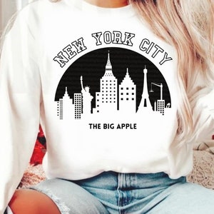 May include: White sweatshirt with a black graphic of the New York City skyline, including the Statue of Liberty, the Empire State Building, and the Chrysler Building. The graphic also includes the text "NEW YORK CITY" and "THE BIG APPLE".