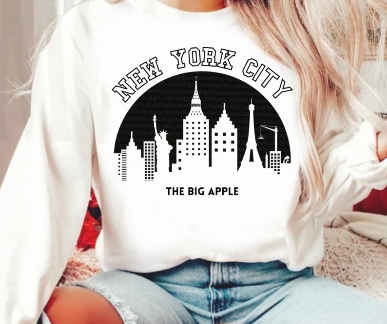 May include: White sweatshirt with a black graphic of the New York City skyline, including the Statue of Liberty, the Empire State Building, and the Chrysler Building. The graphic also includes the text "NEW YORK CITY" and "THE BIG APPLE".