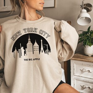 May include: A beige sweatshirt with a black graphic of the New York City skyline and the text "NEW YORK CITY THE BIG APPLE".