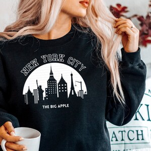 May include: Black sweatshirt with a white graphic of the New York City skyline and the text "New York City" and "The Big Apple".